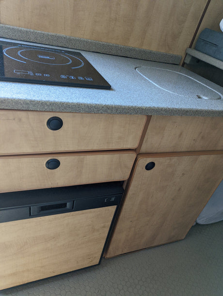 RV rental kitchen: Induction cooktop, covered sink, wood cabinets, and built-in fridge detail.