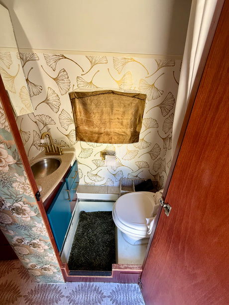 Stylish RV rental bathroom with gold ginkgo leaf wallpaper, blue vanity, sink, toilet, and shower curtain.