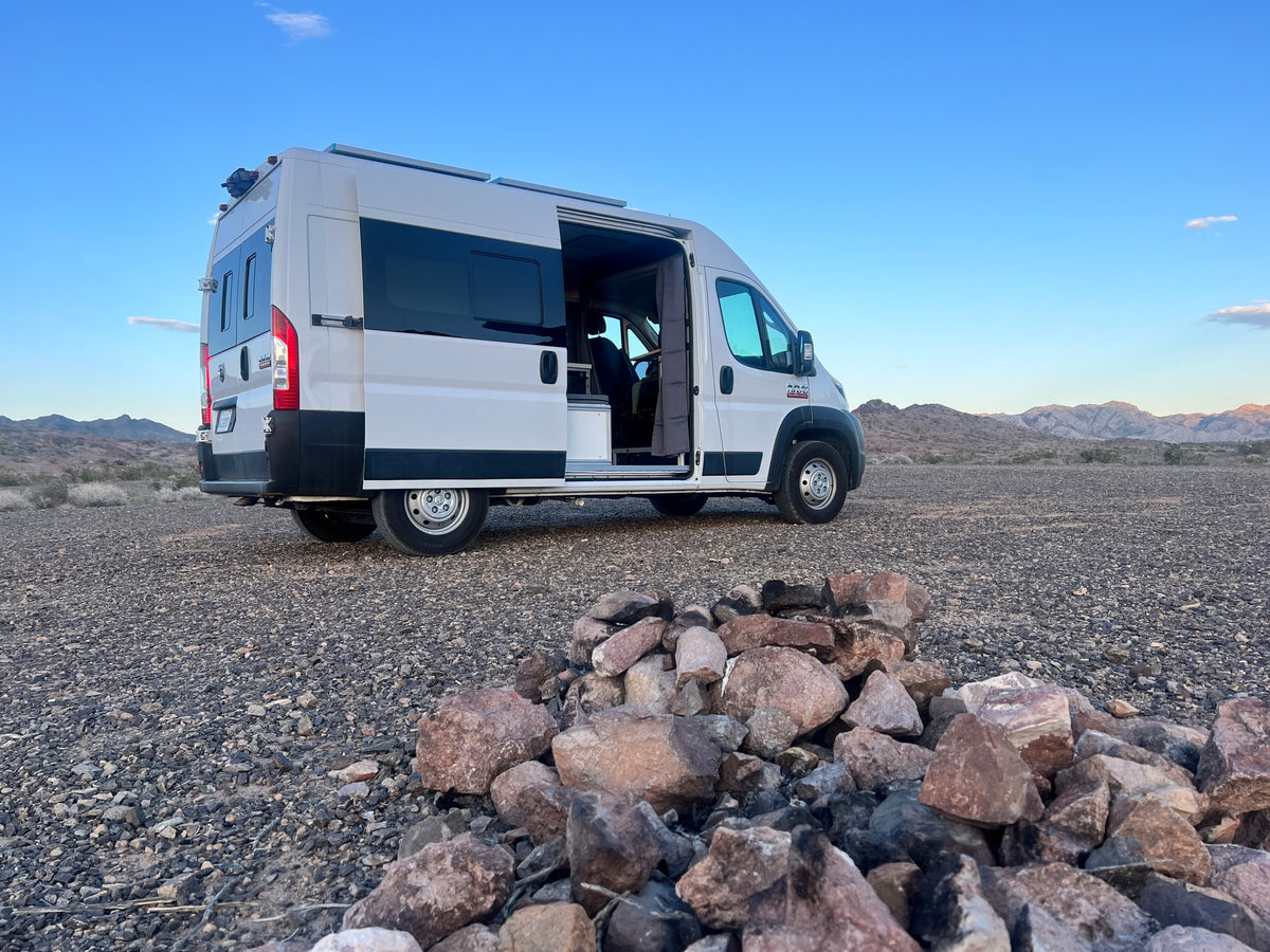 White RV rental van parked in a rocky desert, with mountains under a blue sky and a stone fire pit in the foreground.
