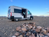 White RV rental van parked in a rocky desert, with mountains under a blue sky and a stone fire pit in the foreground.