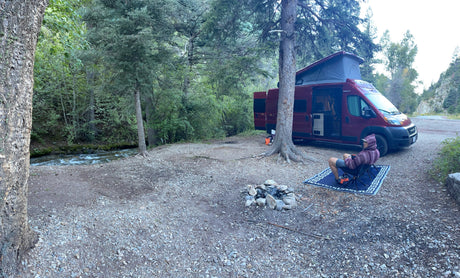 Peaceful RV rental campsite by a river. A person relaxes next to their camper van and fire pit in a scenic forest.