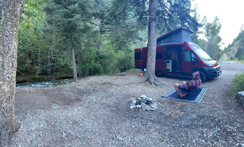 Peaceful RV rental campsite by a river. A person relaxes next to their camper van and fire pit in a scenic forest. — photo 13