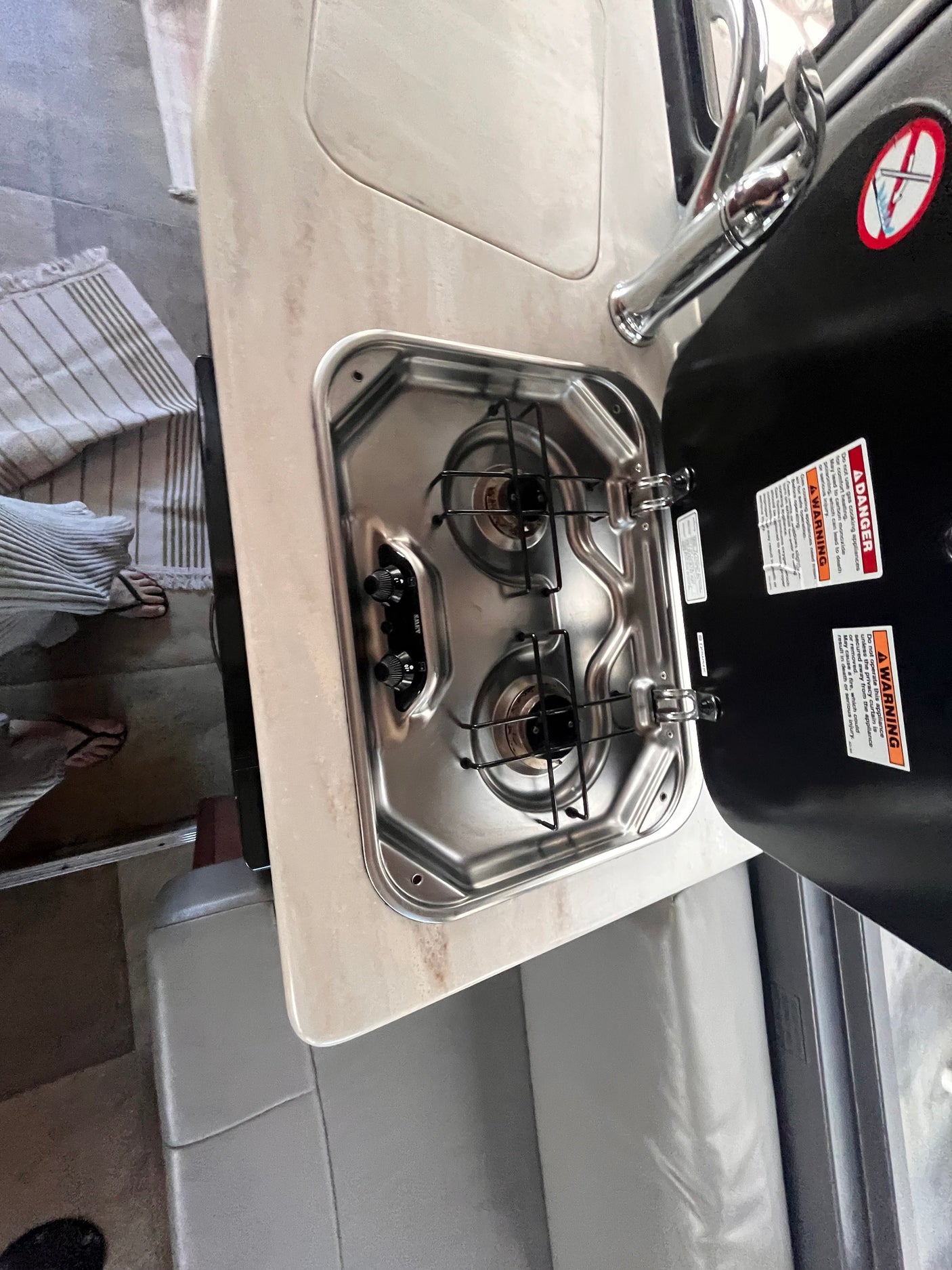 RV rental's compact kitchen with two-burner stove, sink, and faucet. Stove lid open, showing safety warnings.