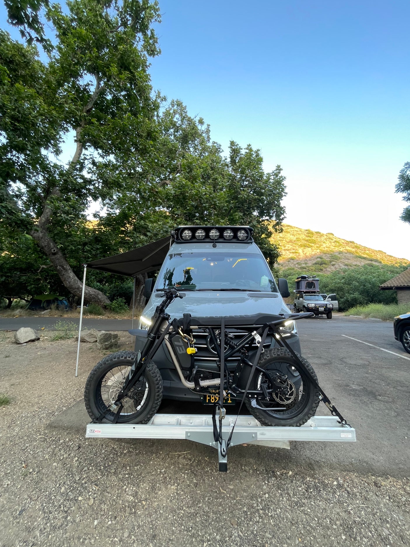 RV rental ready for adventure! Camper van with an electric motorcycle on a hitch carrier at a scenic campsite.