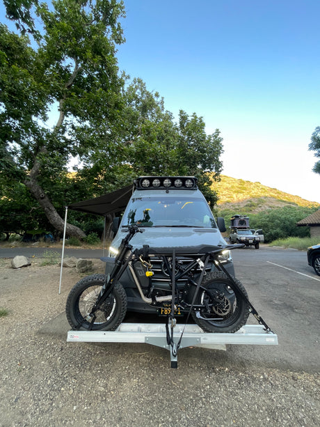 RV rental ready for adventure! Camper van with an electric motorcycle on a hitch carrier at a scenic campsite.