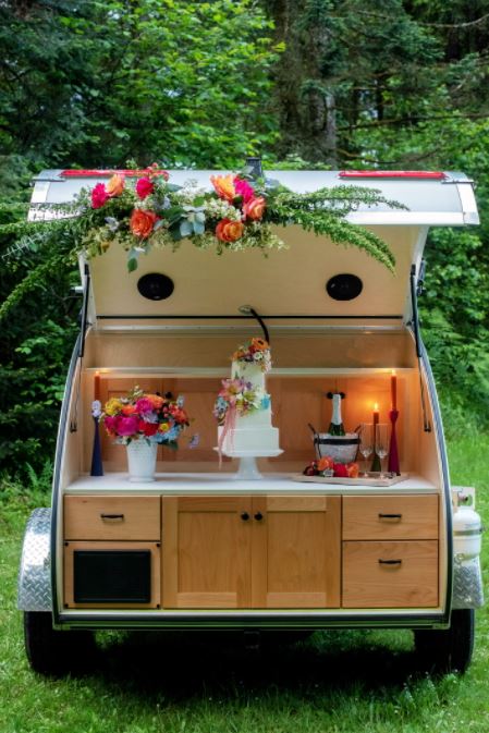 Elegant rv rental teardrop trailer, styled with flowers, presenting a cake and bubbly in its mobile kitchen. Perfect for outdoor events.