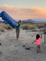RV rental fun: A man and child enjoy a vast desert landscape and sunset, holding blue outdoor gear.