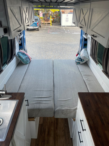 RV rental interior: comfy grey bed, kitchenette, and storage cabinets in a custom camper van. View out back doors.