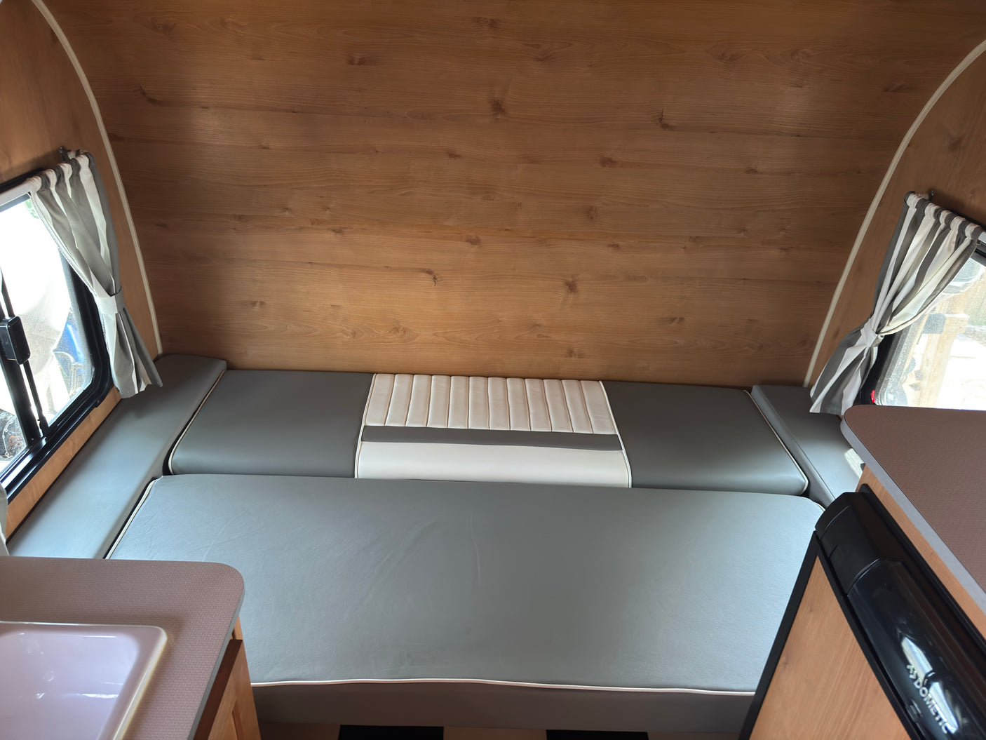 RV rental interior: wood-paneled walls, gray & white convertible bed/seating, striped curtains, sink, & Dometic fridge.