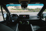 Inside a modern RV rental: Mercedes Sprinter cockpit with a large infotainment screen, steering wheel, and cupholders. Road view ahead.