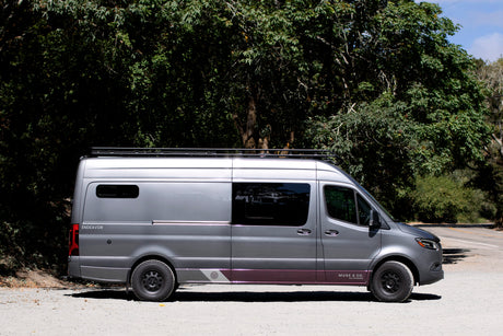 This grey custom Mercedes Sprinter van, an 'Endeavor' rv rental, is parked roadside surrounded by green trees.