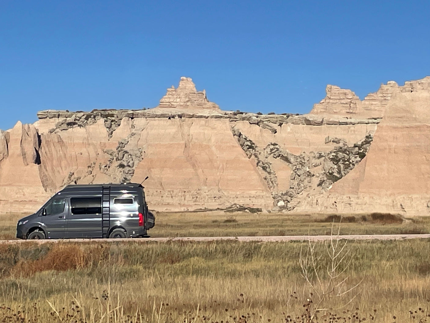 An RV rental drives through a scenic Badlands landscape, featuring unique geological formations under a clear blue sky.