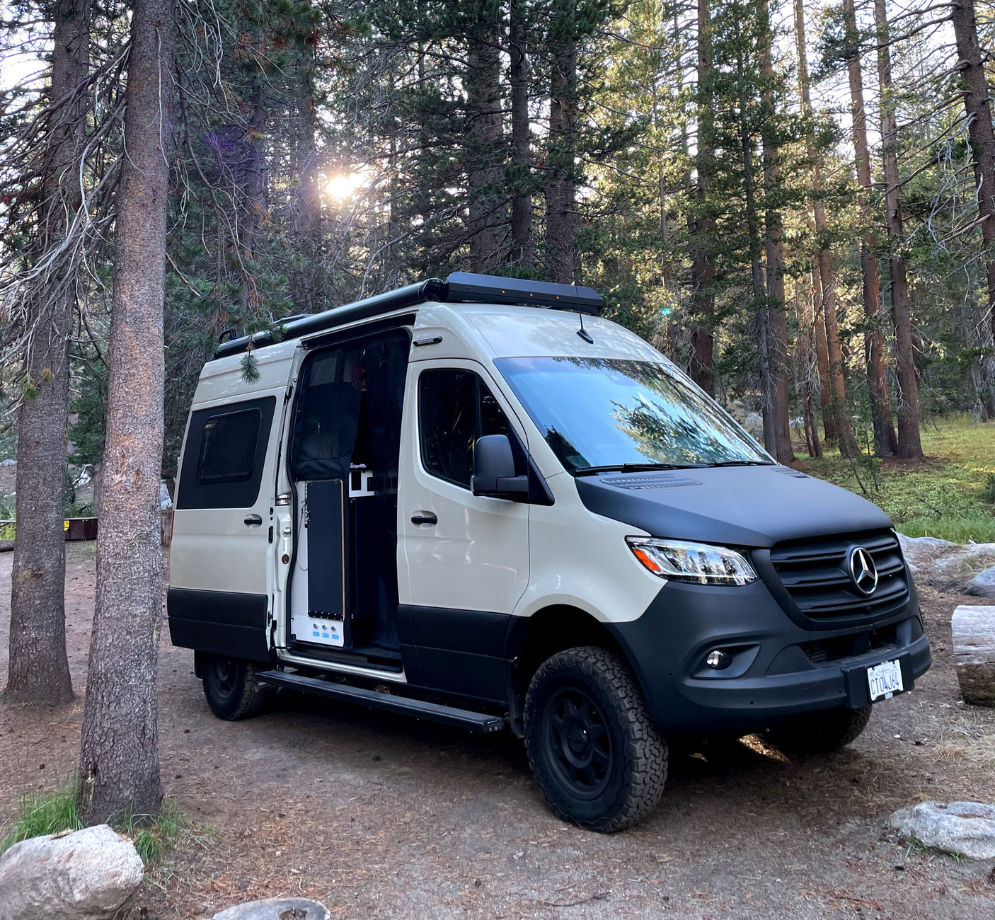 A two-tone Mercedes Sprinter camper van, likely an RV rental, is parked amidst tall trees in a sunny forest.