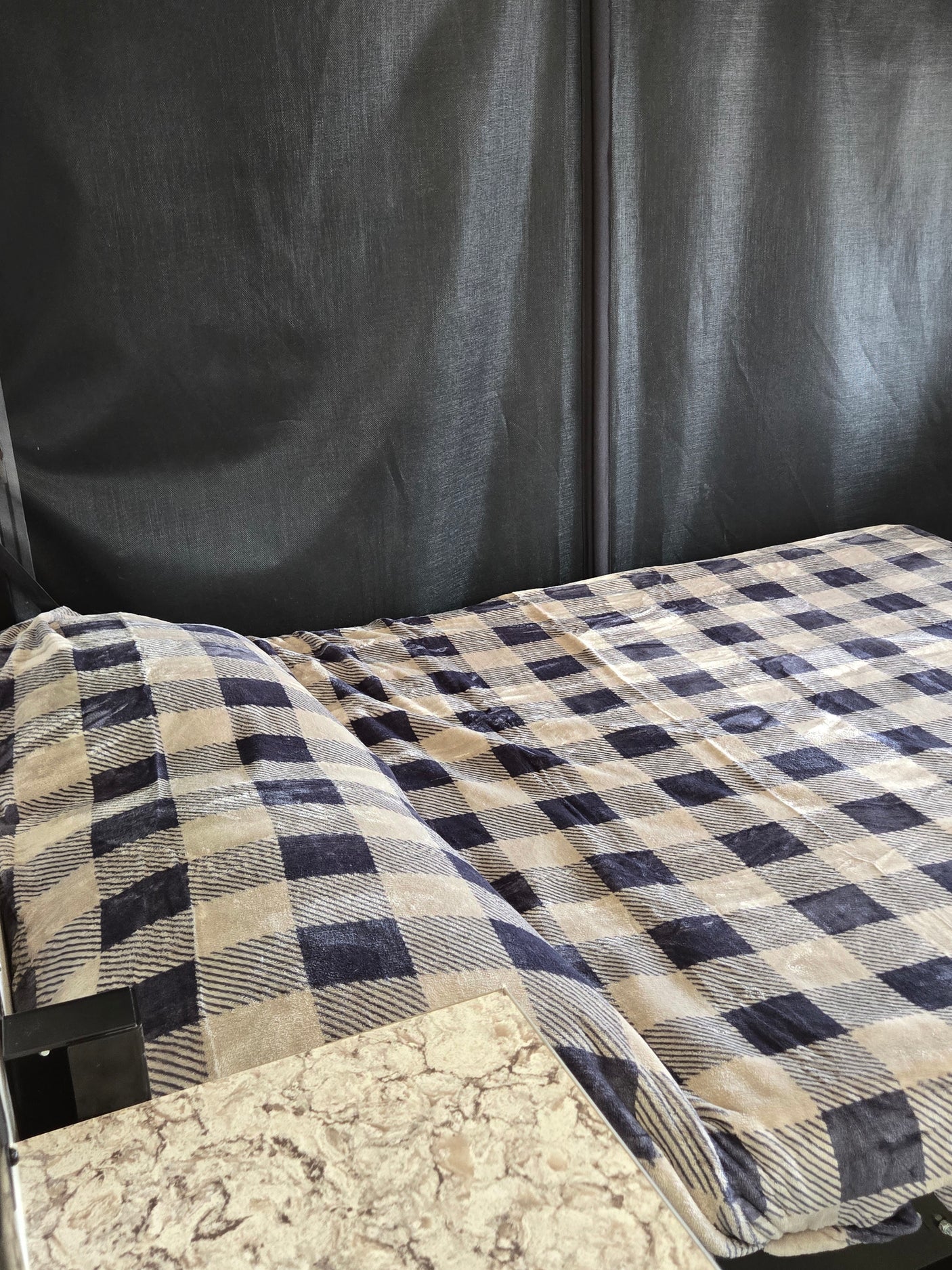 Cozy RV rental interior: A bed with a blue and beige plaid blanket rests against a dark fabric wall, next to a marble-look countertop.