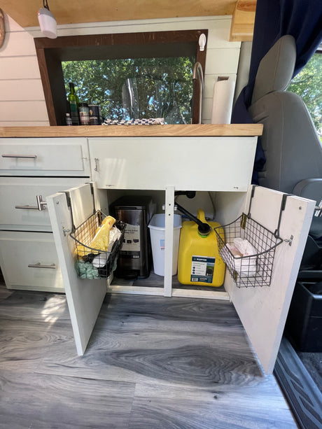 Inside an RV rental, a compact kitchen with open under-sink cabinets revealing organized storage, water tanks, and a trash bin.