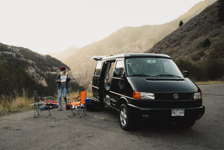 Scenic mountain view with a black VW camper van, chairs, and a woman. Ideal RV rental for your next outdoor adventure.