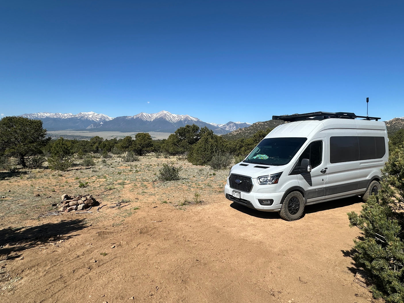 A white rv rental van is parked on a dirt path, with stunning views of snow-capped mountains under a clear blue sky.