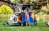 Family enjoying their RV rental: a blue teardrop trailer with a rooftop tent. Girl is inside the tent, boy climbs up, adults relax.