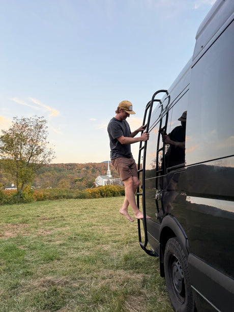 A person climbs a ladder on a black RV rental van, set against a scenic autumn landscape with a distant white church.