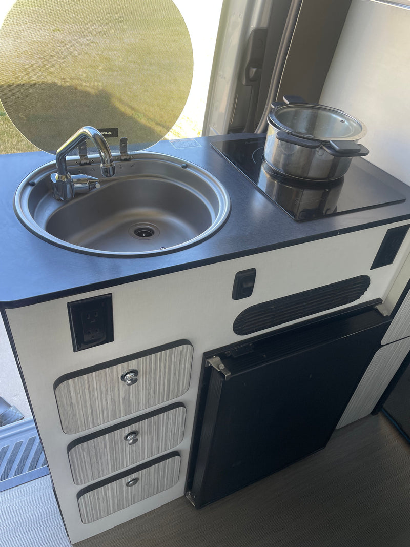 RV rental's compact kitchen features a stainless steel sink, electric cooktop, storage drawers, and under-counter fridge. — photo 11