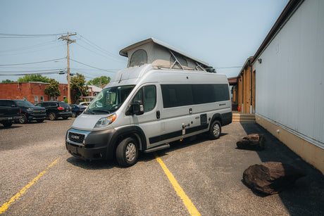 A sleek silver pop-top camper van, an ideal RV rental, parked on a gravel lot with other vehicles and buildings.
