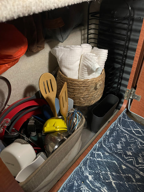 An RV rental's under-cabinet reveals compactly stored kitchen supplies: white towels, wooden utensils, red colanders, and a dish rack.