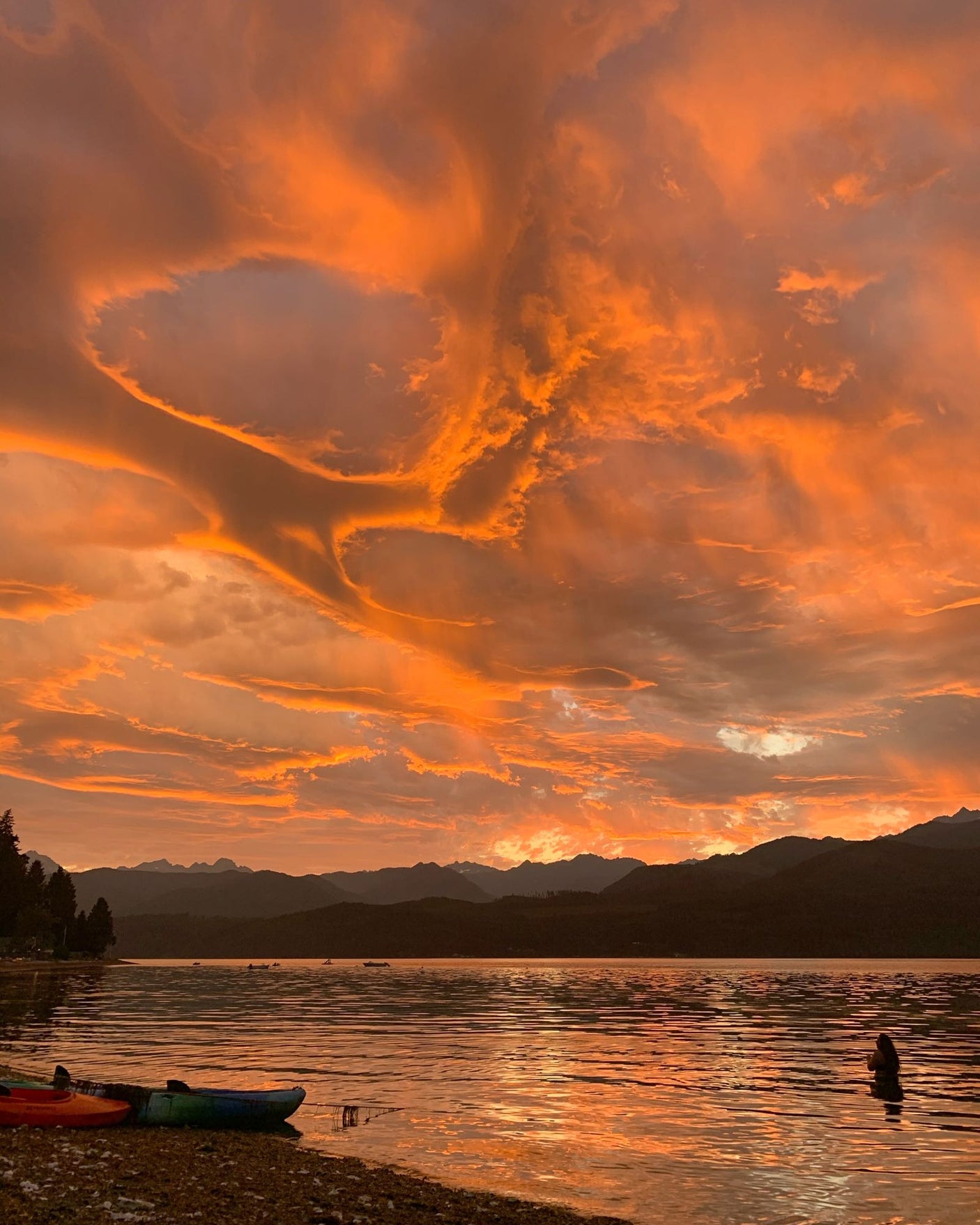 Fiery orange sunset clouds over a mountain lake with kayaks on the shore. A person wades in the water. Book your RV rental getaway!
