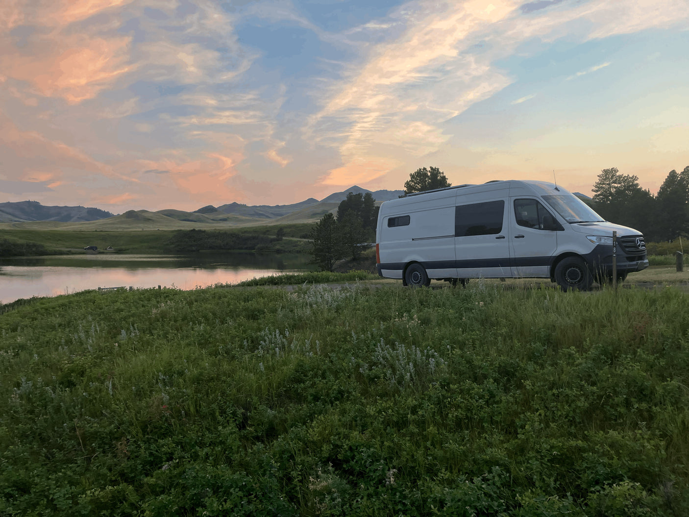 White RV rental parked at a tranquil lake campsite with hills & a vibrant sunset. Perfect nature escape!