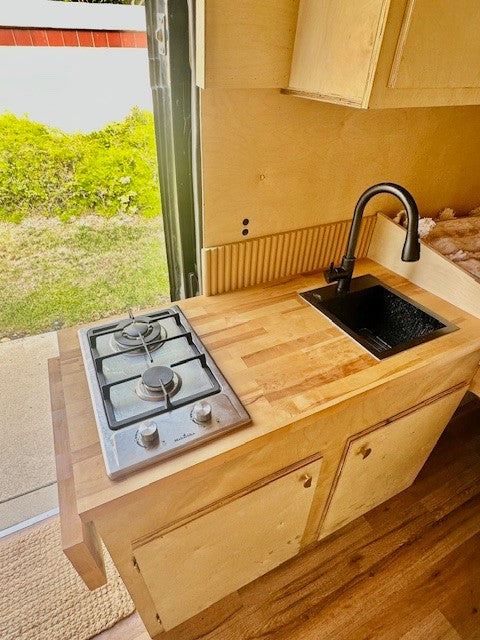Inside an RV rental: a bright, compact kitchen with wooden counters, a two-burner stove, and a modern black sink.