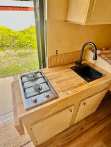 Inside an RV rental: a bright, compact kitchen with wooden counters, a two-burner stove, and a modern black sink.