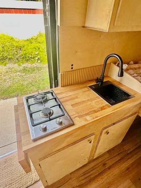 Inside an RV rental: a bright, compact kitchen with wooden counters, a two-burner stove, and a modern black sink.