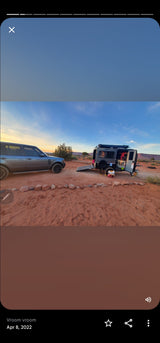 Grey SUV & open teardrop rv rental parked on red desert dirt under a colorful sunset sky.