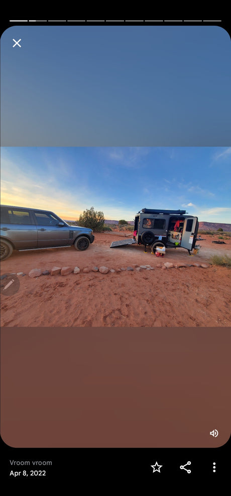 Grey SUV & open teardrop rv rental parked on red desert dirt under a colorful sunset sky.