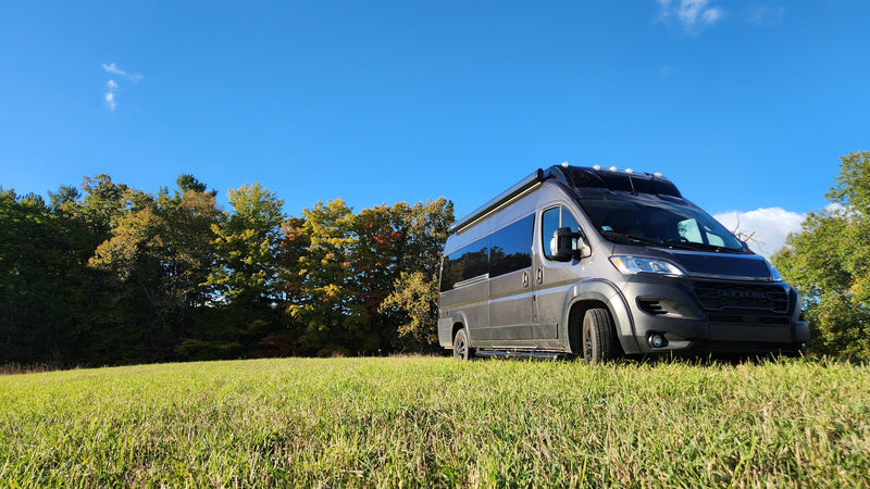 A grey RV rental camper van rests in a sunny green field, surrounded by fall trees under a blue sky. — photo 11