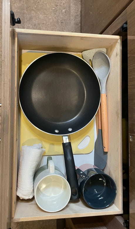 An RV rental kitchen drawer showcases a neatly organized frying pan, utensils, mugs, and cutting board.
