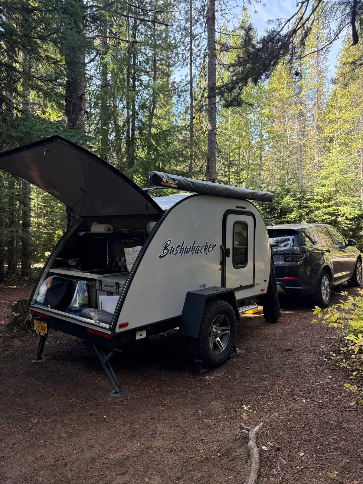 RV rental adventure: Bushwhacker teardrop camper & SUV amidst tall forest trees, kitchen open for camping.