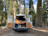 Cozy RV rental camper van with open back doors, bed, and gear, nestled among towering trees in a peaceful forest.