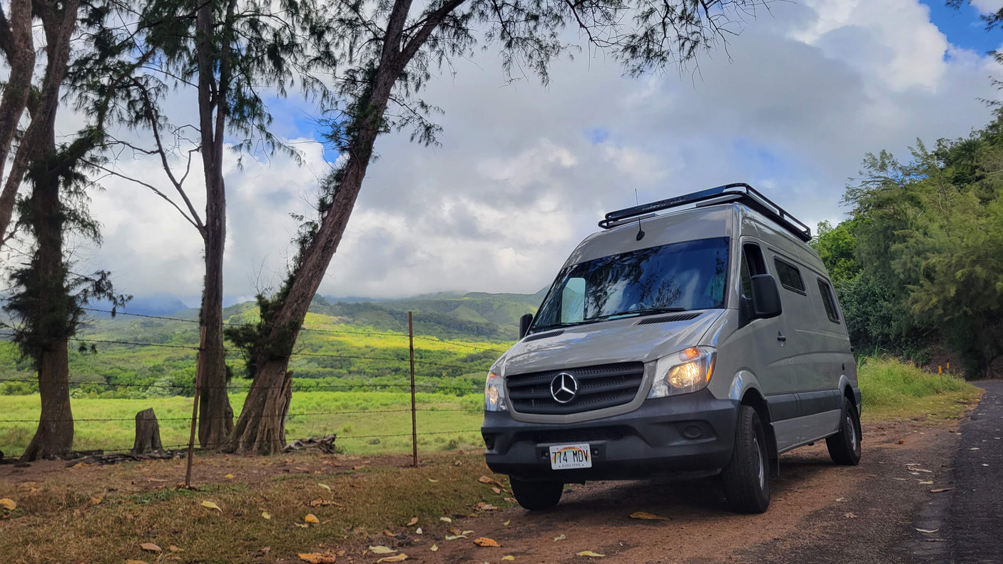 Grey Mercedes Sprinter van RV rental, parked on a dirt path overlooking a lush Hawaiian valley with mountains.