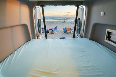 Cozy RV rental bed overlooks a beautiful beach sunset. People relax on sand by ocean waves, fire burning. Perfect beach camping.