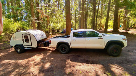 White Toyota Tacoma truck towing a teardrop camper at a sunny forest campsite. Find your ideal RV rental adventure here.