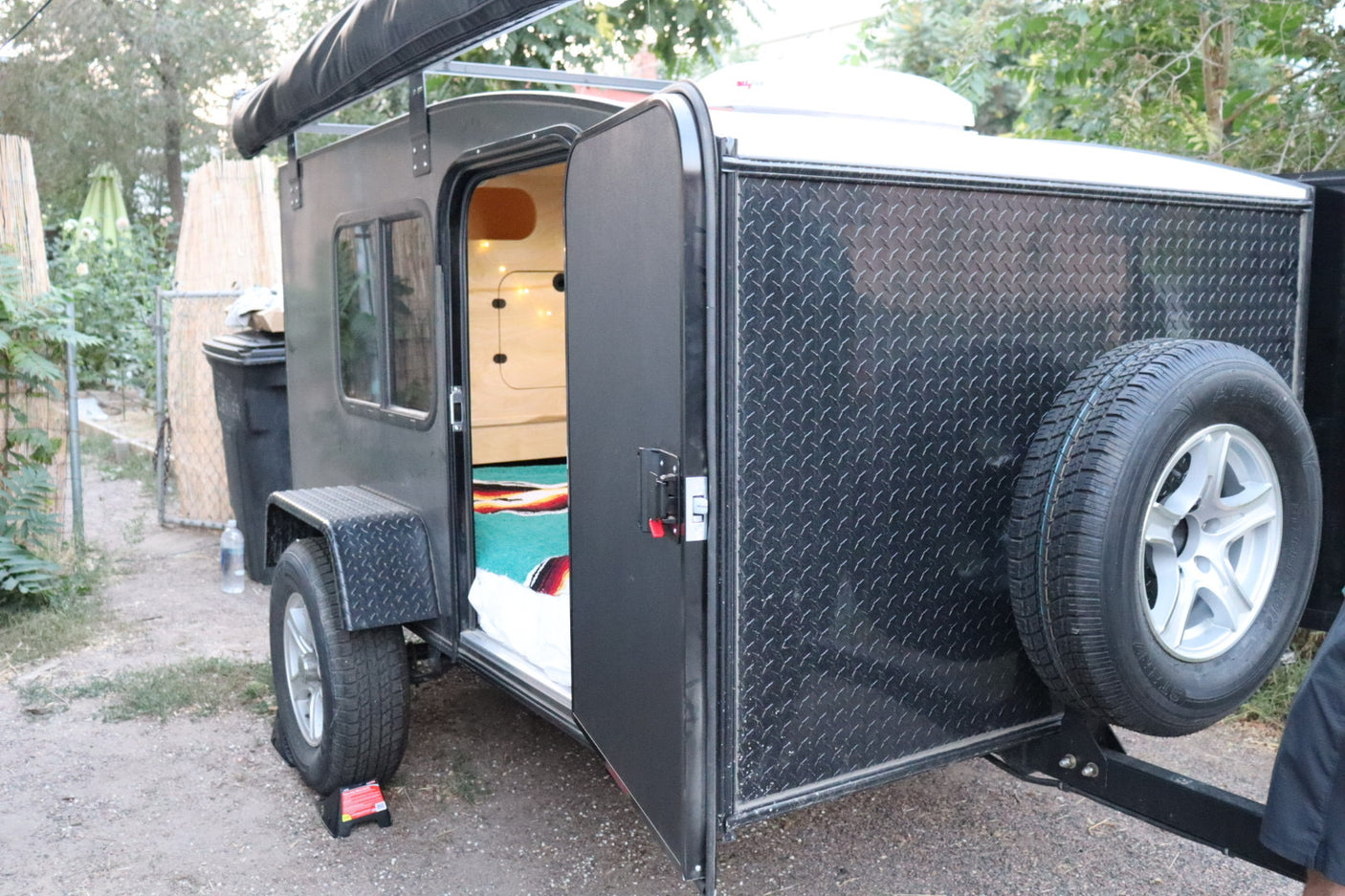Black compact RV rental with open side door revealing a cozy, wood-paneled interior & colorful blanket. Features an awning & spare tire.