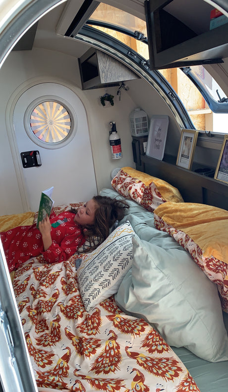 A child reads cozily in bed inside an RV rental. Peacock bedding, a round window, and a compact interior are visible.