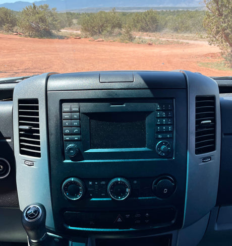 Inside an RV rental: dashboard, Mercedes-Benz gear shift, and dusty console with a scenic dirt road view.