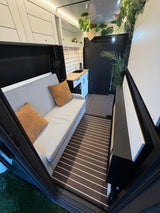This RV rental interior features a modern light gray sofa, compact kitchen with sink, and dark striped floor. Greenery adds charm.