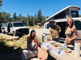 Three friends enjoy meal prep at a sunny campsite with an RV rental and camper vans, embracing outdoor adventures.