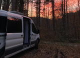 White camper van (rv rental) parked in a rustic forest during a vibrant orange and pink sunset, ideal for nature escapes.
