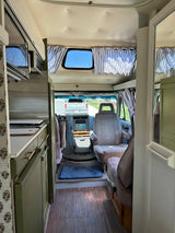 RV rental interior view: kitchenette with olive cabinets, cockpit, front seats, and a truck outside. Cozy camper van.