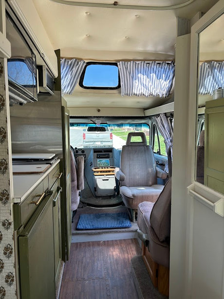 RV rental interior view: kitchenette with olive cabinets, cockpit, front seats, and a truck outside. Cozy camper van.