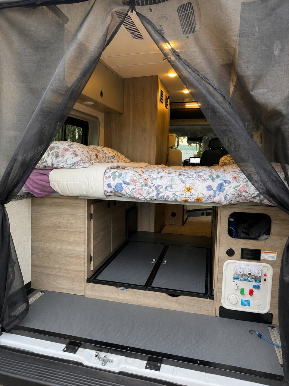 RV rental interior: A bed, storage, and utility connections are visible from the rear, framed by a black mesh screen.
