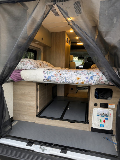RV rental interior: A bed, storage, and utility connections are visible from the rear, framed by a black mesh screen.
